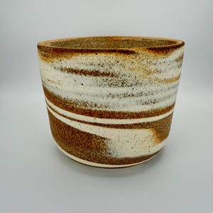 Może przedstawiać: Ceramiczna miska o szerokim, cylindrycznym kształcie. Miska posiada poziome pasy w kolorze brązowym, białym i nakrapianym beżowym. Obręcz jest ciemniejsza brązowa, a wnętrze niewidoczne. Miska jest ustawiona na białym tle.