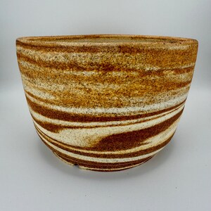 May include: A handcrafted ceramic bowl with a wide, open design. The bowl features a unique striped pattern in shades of brown, tan, and cream, creating a natural, earthy aesthetic. The bowl's dimensions are approximately 10 inches in diameter and 5 inches in height.
