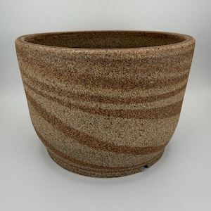 May include: A wide, round ceramic planter with a speckled, textured surface featuring swirling bands of earthy brown and tan tones. The pot has a thick, rounded rim and a slightly tapered base, showcasing a natural, granular clay finish throughout.