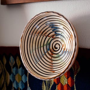May include: A ceramic bowl with a spiral design. The bowl features concentric black lines against a cream background, with accents of blue, orange, and green. The rim is a warm brown color. The bowl is likely handmade and adds a rustic touch.