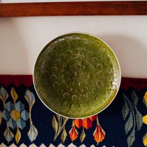 May include: A round ceramic bowl with a glossy olive green glaze and a detailed, embossed floral pattern covering the interior surface. The bowl features a white rim and a textured, repeating circular design that radiates from the center.