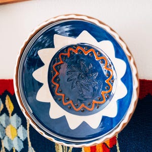 Może przedstawiać: Okrągła ceramiczna miska z głęboką niebieską glazurą i białą, falistą obwódką. W centrum znajduje się wytłoczony wzór liścia paproci otoczony falistą pomarańczową linią i małymi ciemnobrązowymi kropkami, z karbowanym, falistym brzegiem.