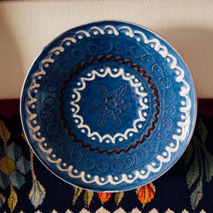 May include: A round ceramic bowl featuring a deep blue glaze with embossed white scrollwork patterns, a central floral motif, and a dark brown wavy line accent. The bowl has a textured surface with repeating circular stamps along the rim.