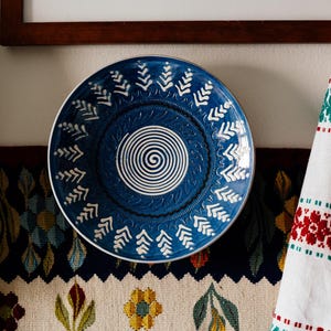 May include: A round, blue decorative plate with a white spiral center and white leaf-like patterns around the rim. The plate is displayed against a wall with a framed artwork and a patterned textile.