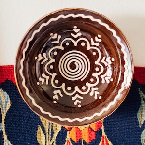 May include: A round, dark brown ceramic bowl featuring a hand-painted white spiral design at the center, surrounded by a scalloped floral pattern and a wavy line along the rim. The bowl has a glossy finish and a smooth, circular shape.