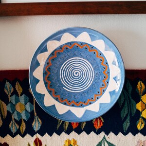 May include: A round ceramic plate featuring a central white spiral on a blue background, surrounded by a ring of small stamped circular patterns and a wavy orange line. The rim is decorated with a white scalloped petal design on blue.