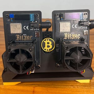 Peut inclure: Deux appareils électroniques noirs avec des ventilateurs, étiquetés "Bitaxe" et "601", sont présentés. Un emblème Bitcoin doré est centré entre eux. Les appareils comportent divers composants, dont un petit écran et un marquage CE. La base est noire avec des accents jaunes.