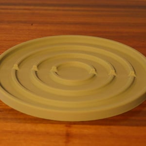 Può includere: Un sottopentola rotondo, color verde oliva chiaro, con scanalature circolari concentriche. Il sottopentola ha un cerchio centrale e tre anelli esterni, ciascuno con piccoli ritagli a forma di freccia. È realizzato in un materiale solido.