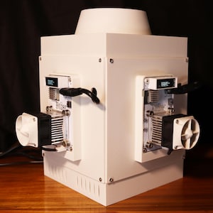 Puede incluir: Un dispositivo electrónico rectangular blanco con una parte superior cilíndrica. Cuenta con dos pequeñas pantallas, ventiladores y el texto "Bitaxe". Se ven cables y tornillos negros. El dispositivo está sobre una superficie de madera.