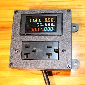 May include: A black electrical device with a digital display showing voltage, current, and frequency readings. Below the display is a black electrical outlet with two receptacles. The device is mounted on a wooden surface.