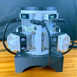 May include: A complex electronic device with the text "The Gamma Core" at the top and "BITAXE 601, 602 and Duo 650" at the bottom. The device has multiple components, including fans, circuit boards, and screens, connected by black cables. It sits on a black base.
