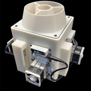 Puede incluir: Un dispositivo blanco impreso en 3D con una parte superior cilíndrica y una base cuadrada. Cuenta con una pequeña pantalla que muestra texto, un ventilador y varios componentes electrónicos. El dispositivo está conectado por cables negros.