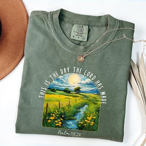 Puede incluir: Camiseta verde oliva con un gráfico de paisaje que representa un río, flores amarillas y un sol. El texto "THIS IS THE DAY THE LORD HAS MADE" está arqueado sobre la escena, con "Psalm 118:24" debajo. Un collar dorado descansa sobre la camiseta.