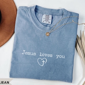 Puede incluir: Una camiseta color azul jean con cuello redondo y mangas cortas. La prenda presenta el texto blanco "Jesus loves you" sobre un icono de corazón blanco que contiene una cruz. Un collar dorado con un pequeño colgante circular descansa sobre ella.