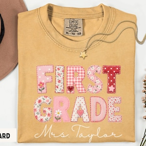 Puede incluir: Una camiseta color amarillo mostaza con el texto FIRST GRADE en estilo patchwork con varios patrones florales y de cuadros en rosa, rojo y blanco. Debajo del texto, el nombre Mrs. Taylor está impreso en letra cursiva blanca. Cuello redondo.