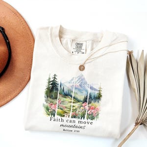 Puede incluir: Camiseta color crema con un gráfico de montaña y flores. El diseño incluye una cordillera, árboles y flores, con el texto "Faith can move mountains" y Mateo 17:20. También se ven un collar dorado y un sombrero marrón.