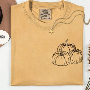 Puede incluir: Una camiseta de manga corta color amarillo mostaza con un gráfico de líneas negras de tres calabazas en el pecho. La prenda tiene cuello redondo y una etiqueta de la marca Comfort Colors en el interior. El texto MASTURD es visible a la izquierda.