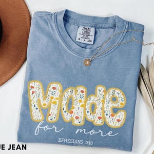 Puede incluir: Una camiseta azul vaquera Comfort Colors con las palabras "Made for more" en un diseño floral. La camiseta también tiene el texto "Efesios 2:10" y un collar dorado con un colgante.