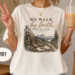 Puede incluir: Una camiseta de color marfil con un estampado gráfico que incluye el texto WE WALK by faith NOT BY SIGHT 2 Corinthians 5:7. La ilustración muestra una señal de madera con la palabra FAITH, una mochila, botas de montaña, una brújula y montañas.