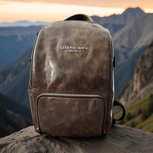 May include: A brown leather backpack with the text "ETERNAL ROCK CHICAGO" in silver lettering. The backpack has a front zippered pocket and a top handle. The background features a mountain range.