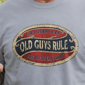 May include: A light gray t-shirt with the text "OLD GUYS RULE" in an oval design. The design is in red, black, and gold. The shirt is worn by a person wearing a black baseball cap.