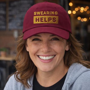 May include: A maroon baseball cap featuring the gold embroidered words "SWEARING HELPS". The cap is worn by a person smiling. The background is blurred, with out-of-focus lights.