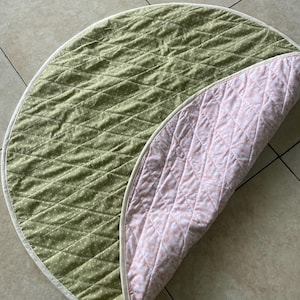 May include: A reversible, quilted play mat. One side is olive green with white polka dots, the other is pink with a white floral pattern. The mat has a white border and a diamond-stitched pattern.