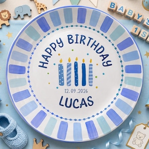 May include: A round white ceramic plate featuring a blue striped border and a central design of five blue birthday candles. The text reads HAPPY BIRTHDAY, the date 12.09.2026, and the name LUCAS in dark blue lettering with small colorful dots.