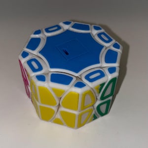 May include: A multi-colored puzzle cube with a unique shape. The cube features blue, yellow, white, green, and pink sections. The puzzle has a complex design with various geometric shapes and patterns, offering a challenging brain teaser.