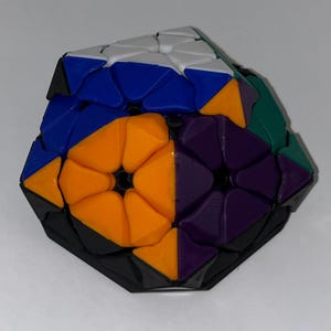 May include: A multi-faceted puzzle cube featuring a complex geometric design with a black base. The surface is composed of triangular and diamond-shaped segments in vibrant colors including white, blue, orange, purple, and teal, arranged in a pattern.