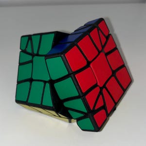 May include: A multi-colored puzzle cube with a unique, non-cubical shape. The puzzle features red, green, blue, and yellow panels, outlined in black. The cube is partially solved, with some panels still misaligned.