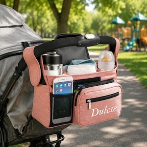 May include: A coral-colored stroller organizer bag attached to a stroller handle. It features a mesh pocket holding a smartphone, a zippered front pocket with the name Dulcie printed in white, and open compartments containing a bottle and a cup.