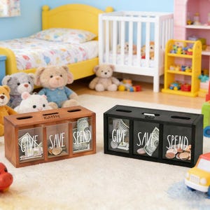 May include: Two wooden and black money boxes with three compartments labeled "GIVE", "SAVE", and "SPEND". The wooden box has the name "Kathy" on top. The compartments are filled with money. The boxes are in a child's bedroom.