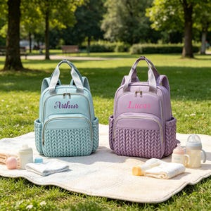 May include: Two baby diaper backpacks, one light blue and one lavender, sit on a white blanket with baby supplies. The light blue backpack has the name "Arthur" embroidered on it, and the lavender backpack has the name "Lucas". Both have a woven pattern on the front.