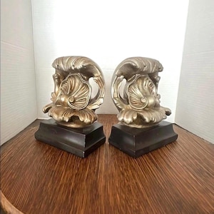 May include: Two decorative bookends with a silver and gold finish. Each bookend features a stylized design with a seashell and wave motif, mounted on a black rectangular base. The bookends are displayed on a wooden surface.