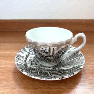 May include: A white teacup and saucer set with a black and white pastoral scene design. The teacup has a curved handle and a scalloped rim. The saucer has a matching design. The set is placed on a wooden surface.