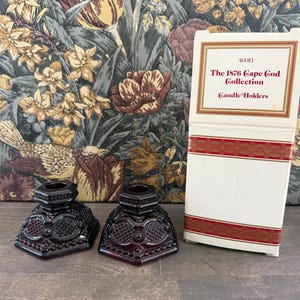May include: Two deep red glass candle holders with a faceted, geometric pattern and a matching white cardboard box. The box features red and gold decorative bands and text that reads: Avon, The 1876 Cape Cod Collection, Candle Holders.