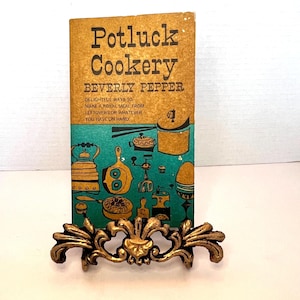 May include: A vintage cookbook titled "Potluck Cookery" by Beverly Pepper, displayed on a decorative gold-colored metal stand. The book has a brown cover with black text and a teal and gold illustration. The stand has an ornate, floral design.