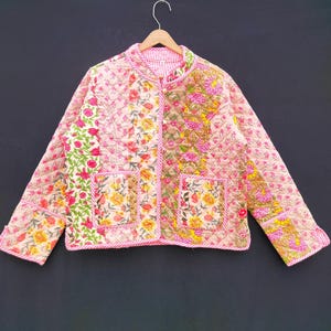 May include: A quilted jacket with a floral pattern in pink, yellow, and green. The jacket has a round collar, two pockets, and a striped lining. The jacket is hanging on a wooden hanger.