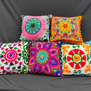 May include: Five square cushions with detailed embroidered patterns. Each cushion displays a unique colour palette, including pink, orange, and blue, with colourful pom-pom edging. The cushions feature floral and geometric designs.