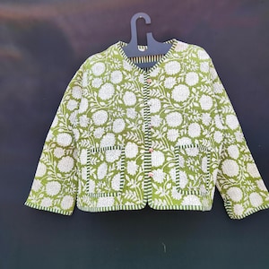 May include: A green quilted jacket with a white floral pattern. The jacket has two front pockets and is buttoned up the front with pink buttons. The jacket is hanging on a black hanger against a dark background.