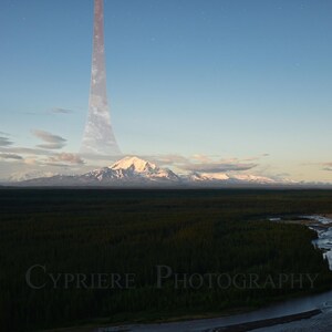 以下が含まれることがあります： 青い空の下、雪をかぶった山々が連なる風景写真。手前には森の中を流れる川があります。空には大きな透明なリング状の構造物があります。「Cypriere Photography」の文字が下にあります。