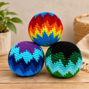 Puede incluir: Tres bolas de ganchillo esféricas con intrincados patrones en zigzag. La bola superior muestra un degradado de arcoíris vibrante, la bola inferior izquierda tonos morados y azules, y la inferior derecha tonos negros, turquesas y verdes.