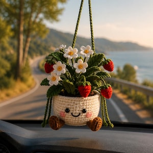 May include: A crocheted hanging plant ornament featuring a cream-coloured pot with a smiling face, pink cheeks, and brown feet. The plant has green leaves, white flowers with yellow centres, and small red strawberries, all suspended by green yarn.