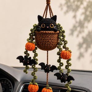 Peut inclure: Un ornement en crochet à suspendre au rétroviseur de voiture représentant un chat noir dans un panier marron. Des vignes vertes pendent du panier, ornées de petites citrouilles orange et de chauves-souris noires, le tout suspendu par des cordons tressés.