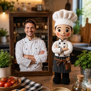 May include: A crochet doll dressed as a chef, wearing a white toque, a white double-breasted jacket with black buttons, a black and white striped apron with a small pocket containing wooden utensils, and black trousers, standing next to a framed photo.