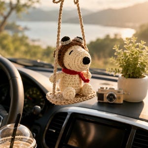 May include: A crocheted cream-coloured dog figure wearing a brown aviator hat with black goggles and a red scarf, sitting on a small crocheted swing suspended by two braided cords, designed to hang from a vehicle rearview mirror.