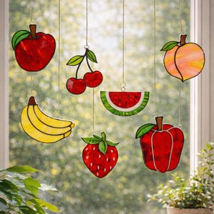 May include: Stained glass fruit ornaments, including a red apple, cherries, a banana, watermelon slice, strawberry, and a peach, hang from a chain. The colorful fruit decorations are suspended in front of a window with a blurred green background.