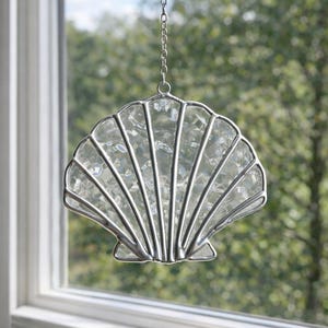 Op de afbeelding: Een hangende schelpvormige suncatcher met een zilverkleurig metalen frame en heldere, getextureerde glazen segmenten. Het ornament in de vorm van een schelp heeft stralende metalen ribben en hangt aan een bijpassende zilverkleurige ketting.