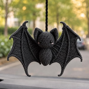 May include: A dark gray crocheted bat hanging from a black braided cord. The bat features large, textured wings, pointed ears, and two small black eyes with tiny pink accents on the cheeks. The yarn construction shows visible stitches throughout.